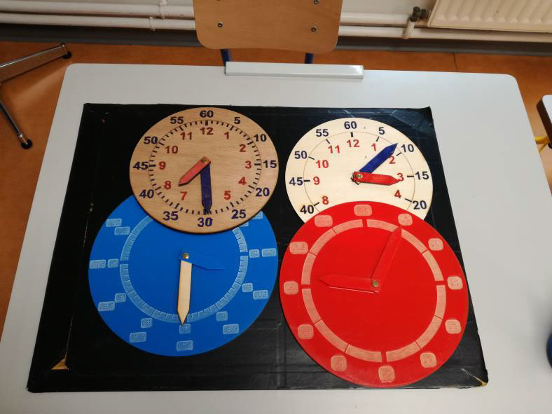 Photo montrant 4 horloges à manipuler, 2 en gros K (bois) et 2 en braille (plexiglas). La numérotation (heures et/ou minutes) et le nombre de graduations varient d'un modèle à l'autre. 